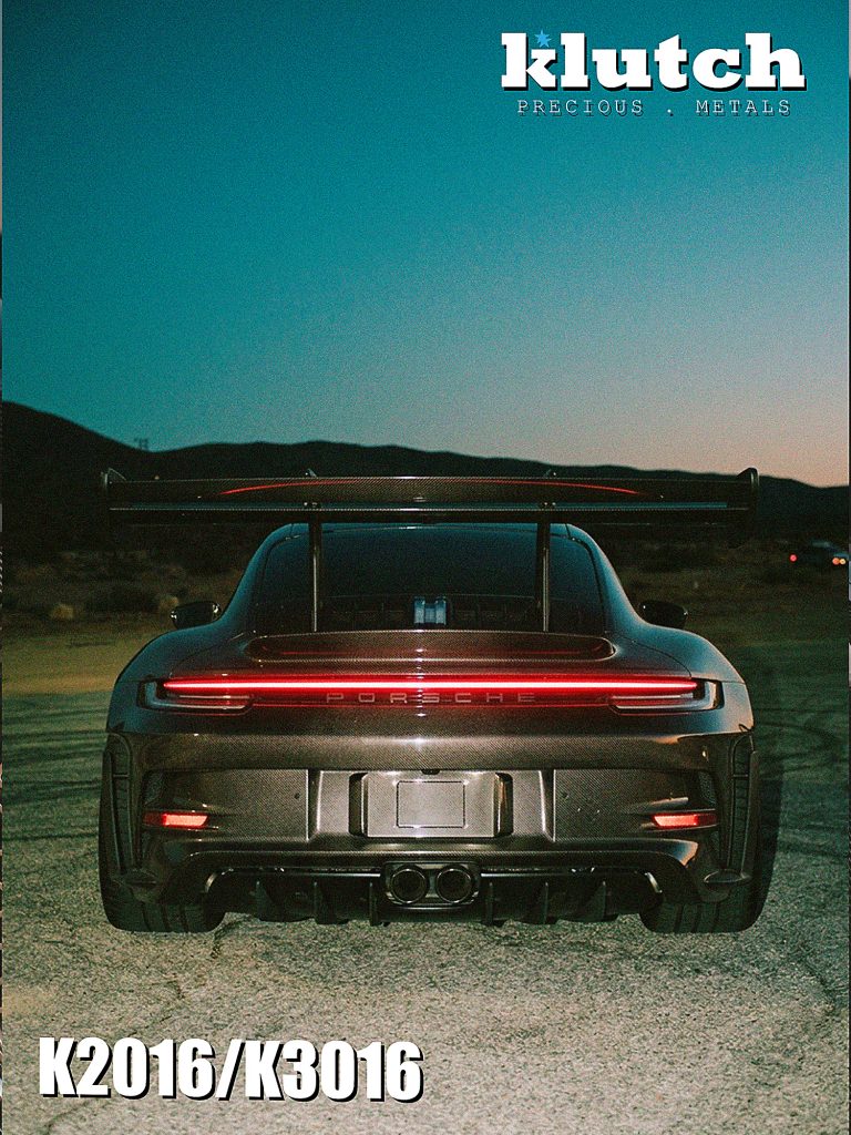 Rear view of a carbon-fiber Porsche 911 with a large rear wing and illuminated taillight at dusk