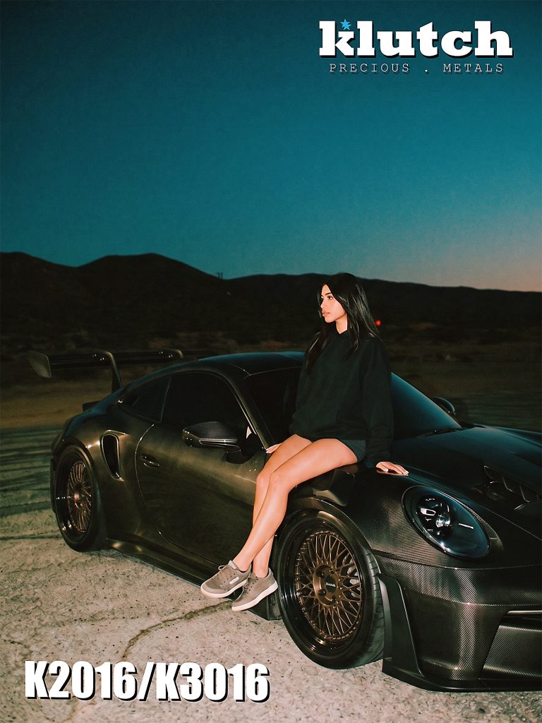 A woman seated on the front fender of a carbon-fiber Porsche 911 fitted with bronze-toned Klutch mesh wheels